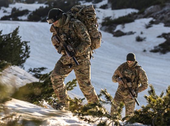 Two fully equipped operators in winter tactical clothing ascending a snowy hillside on a sunny winter day.