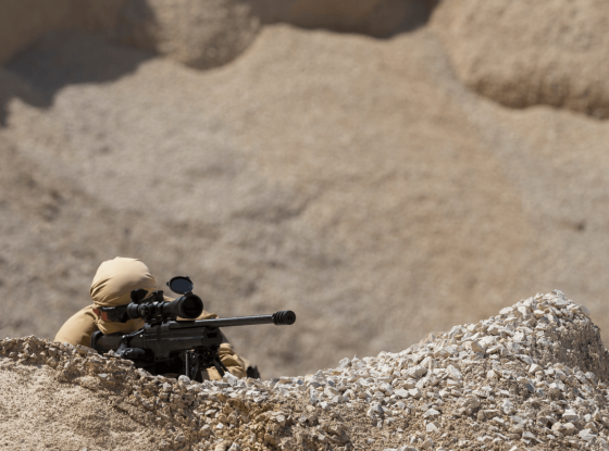 A person in beige tactical gear aims a sniper rifle in a desert-like environment, blending into the arid surroundings for camouflage.