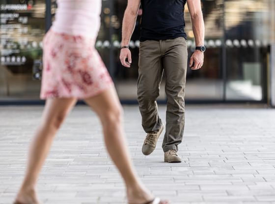 A woman walking in the foreground while a man in EDC tactical clothing walks behind her in an outdoor urban setting, illustrating a potential scenario of being followed.