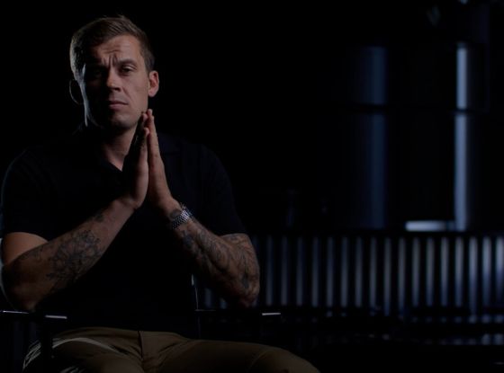 Man seated in a dark room with hands clasped, appearing reflective and focused, symbolizing contemplation of situational awareness.