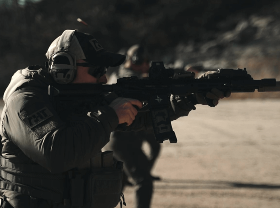 A person wearing tactical gear and headphones aims a rifle outdoors on a dirt surface with another similarly dressed individual visible in the background.